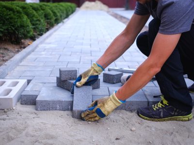 The master in yellow gloves lays paving stones The master in yellow gloves lays paving stones in layers. Garden brick pathway paving by professional paver worker. Laying gray concrete paving slabs in house courtyard on sand foundation base.